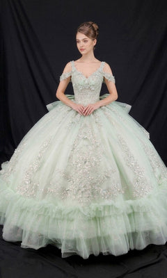 A woman stands in front of a black backdrop wearing the Quinceanera Dress CHF3136 By Chicas in sage, featuring a ballgown skirt adorned with 3D floral appliques and delicate ruffled layers.