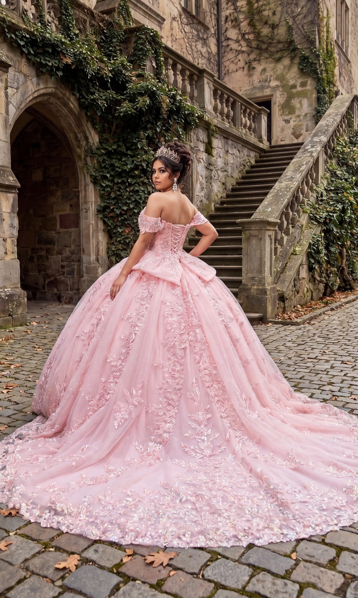 A woman in the Quinceanera Dress CHF3138 By Chicas, an elaborate pink tulle ballgown, stands on a cobblestone path beside ivy-covered stone stairs and a historic building.