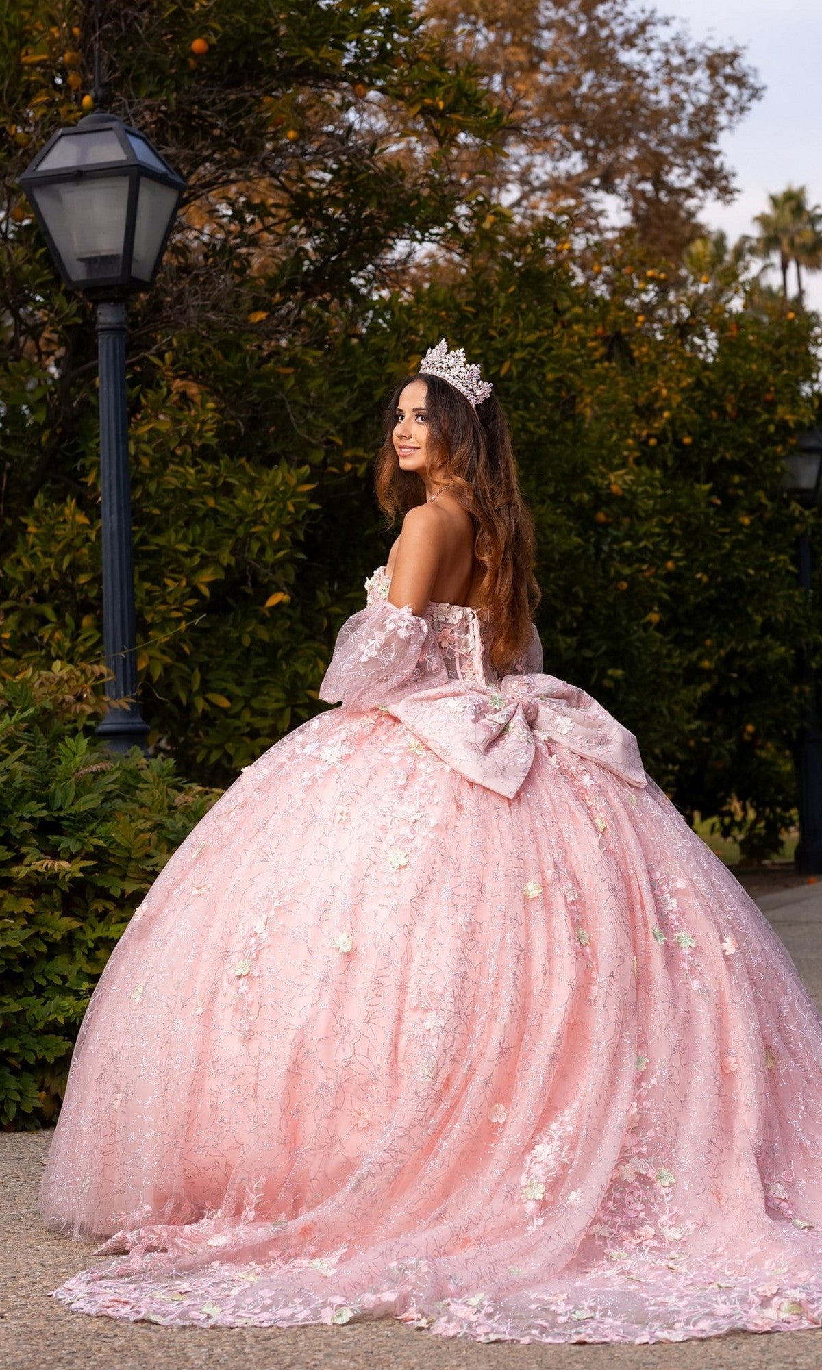 Wearing the Quinceanera Dress CHF3140 by Chicas, a woman stands outdoors in a voluminous pastel ballgown adorned with delicate 3D floral appliques, glancing back over her shoulder as her tiara sparkles in the light.