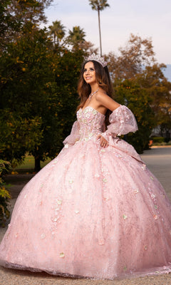 A young woman outdoors stands on a path surrounded by greenery and trees, wearing the Quinceanera Dress CHF3140 by Chicas—a sparkling pink ballgown skirt adorned with 3D floral appliques—and a tiara.