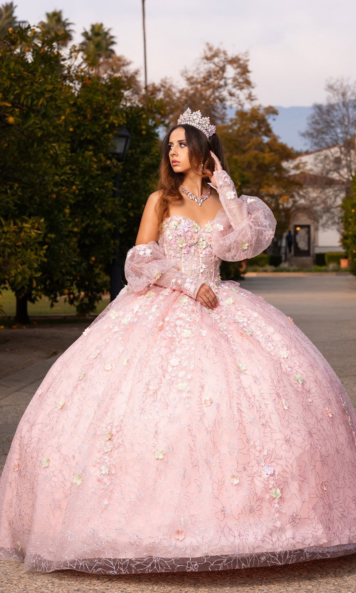 A woman wearing the Quinceanera Dress CHF3140 by Chicas in blush, adorned with 3D floral appliques and a sparkling tiara, stands outdoors looking to the side. The elegant ballgown skirt flows gracefully around her, accentuating the delicate floral details of her ensemble.