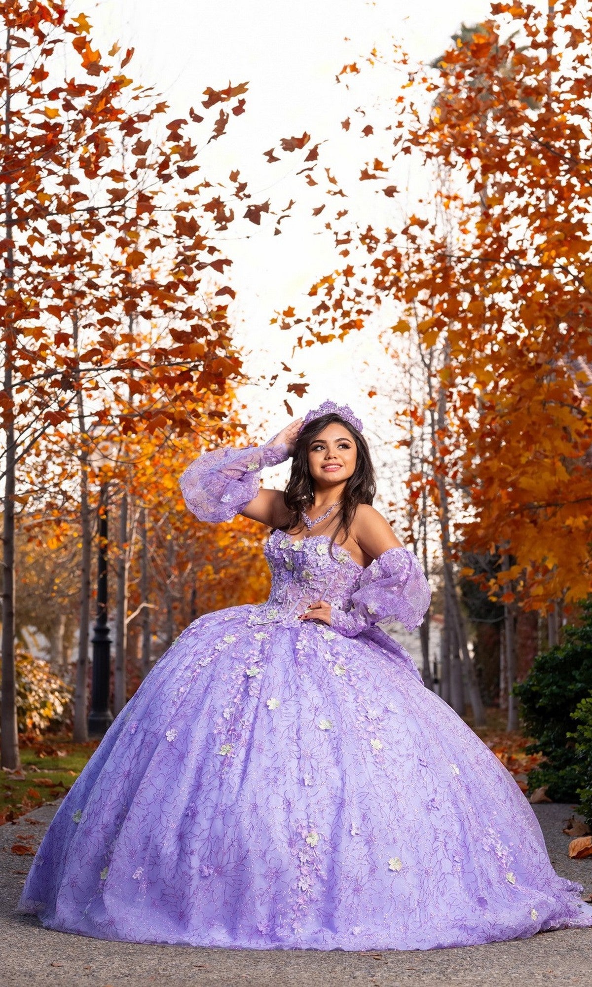 A young woman smiles as she poses on a path lined with autumn trees, wearing the Quinceanera Dress CHF3140 by Chicas in a lovely lilac shade. The dramatic ballgown skirt and delicate 3D floral appliques add elegance to her look as she stands with one hand on her waist and the other adjusting her crown.
