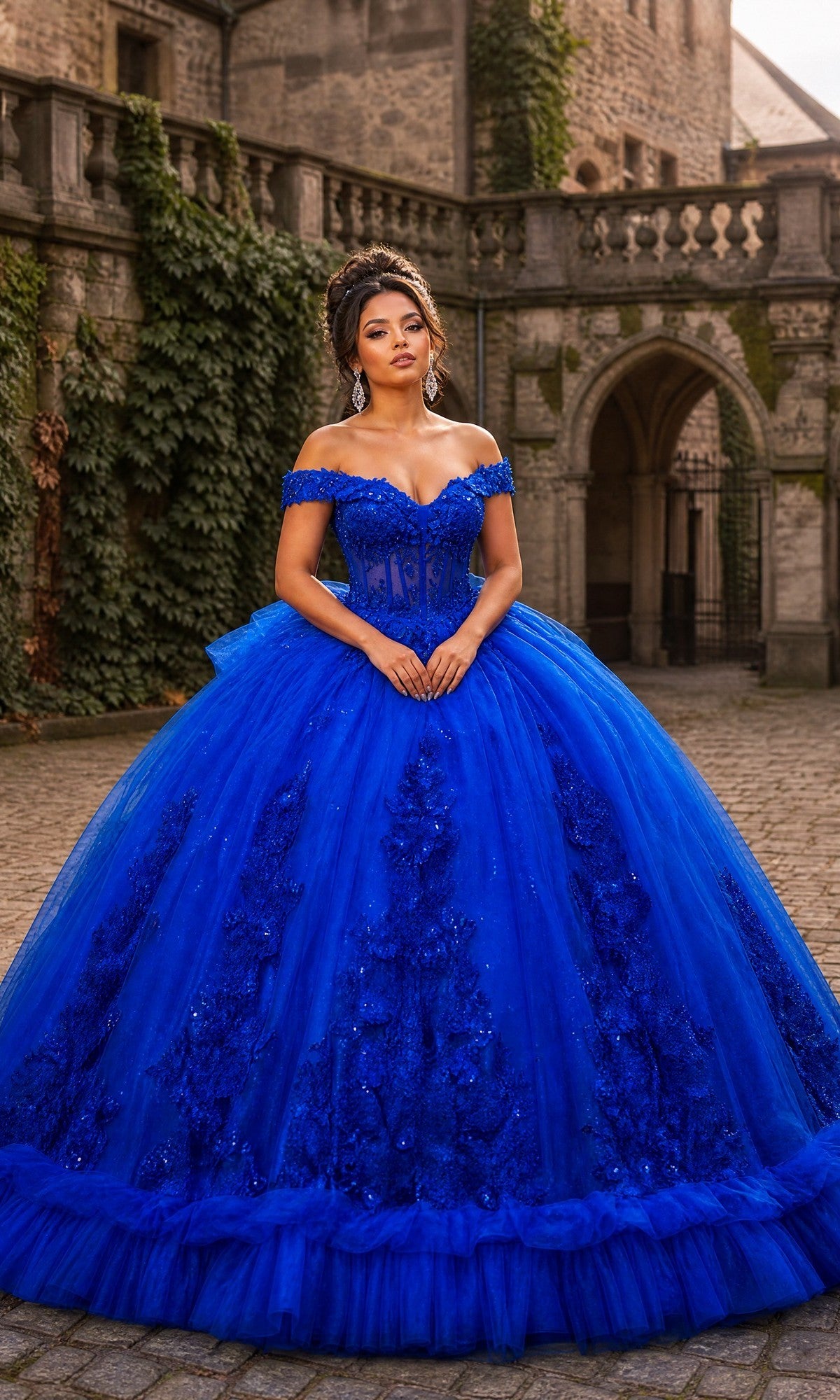 A woman stands on a cobblestone path before an ivy-clad stone castle, wearing the Quinceanera Dress CHF3141 By Chicas in royal blue, featuring an off-shoulder design adorned with delicate 3D floral accents.