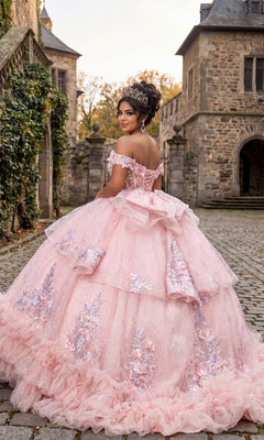A young woman wearing the Quinceanera Dress CHF3141 by Chicas, featuring a pink, ornate design with a flowing ballgown skirt and corset bodice, stands on a cobblestone path outside a stone building and turns to smile at the camera.