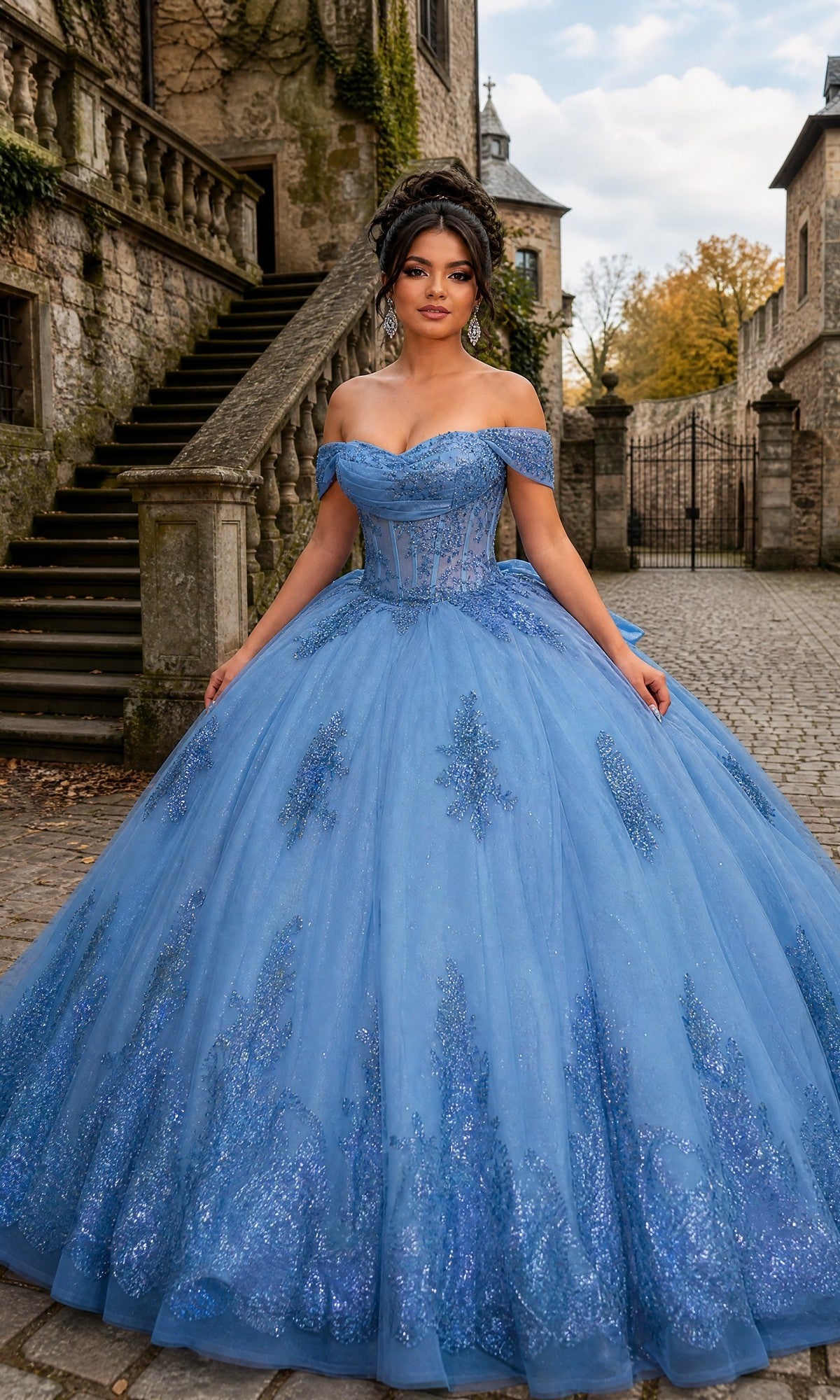 A woman in the Quinceanera Dress CHF3142 By Chicas, a slate blue off-the-shoulder ballgown skirt with glitter details, stands on a cobblestone path near stone steps and historic buildings.