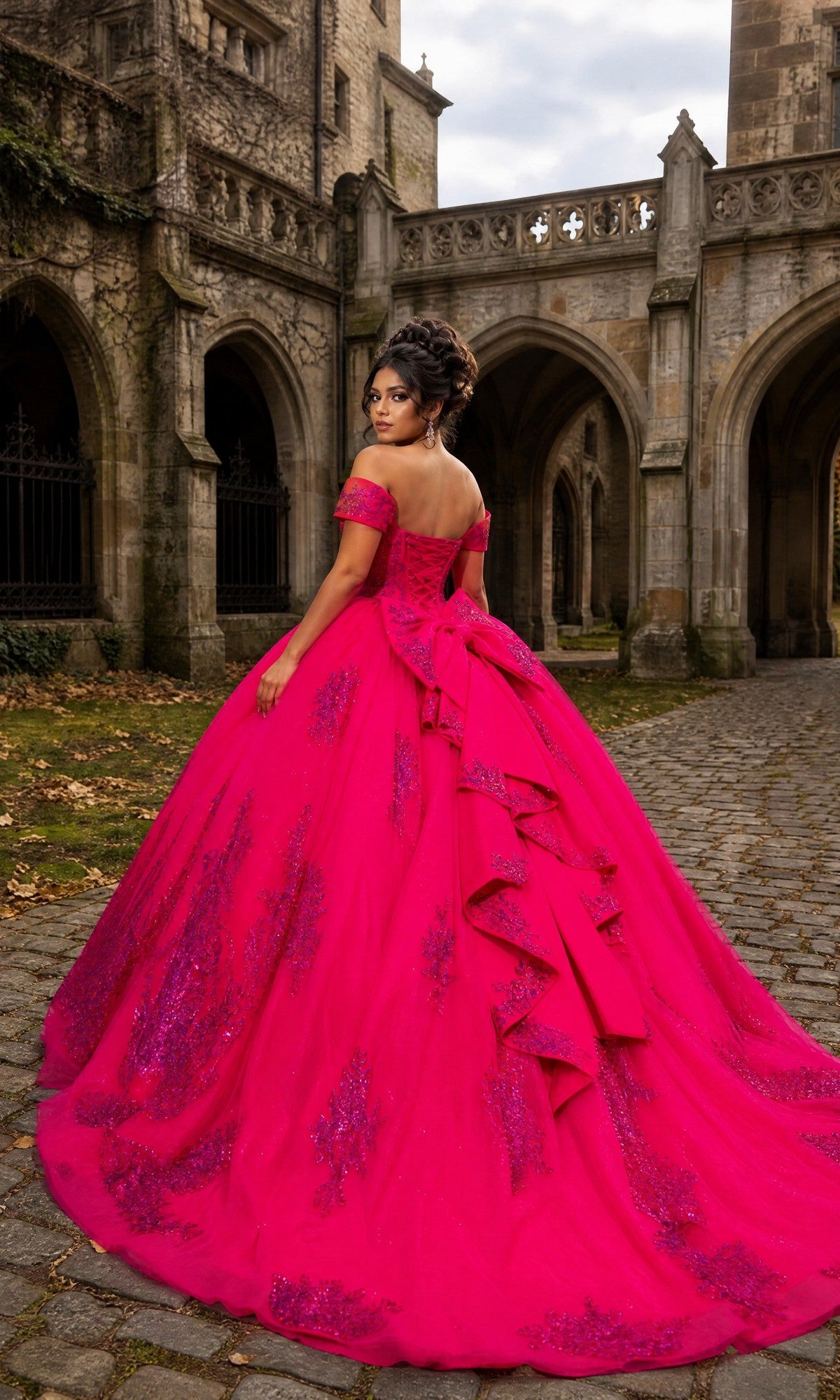 A woman stands on a cobblestone path outside a stone medieval-style building, facing away, wearing the Quinceanera Dress CHF3142 By Chicas – an ornate hot pink gown with a full ballgown skirt.