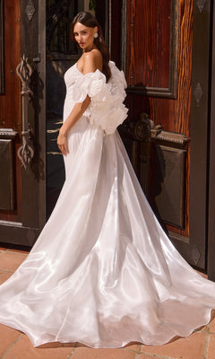 A woman stands in front of wooden doors wearing the Ladivine CJ933W long prom dress, featuring a white off-shoulder design with a mermaid skirt, long train, sweetheart neckline, and large floral accents.