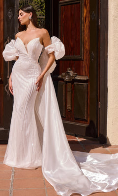 A woman wearing the Ladivine CJ933W long prom dress in off white, featuring a fitted silhouette with a mermaid skirt, deep V neckline, sheer puff sleeves, and a sweeping train, stands by ornate wooden doors.