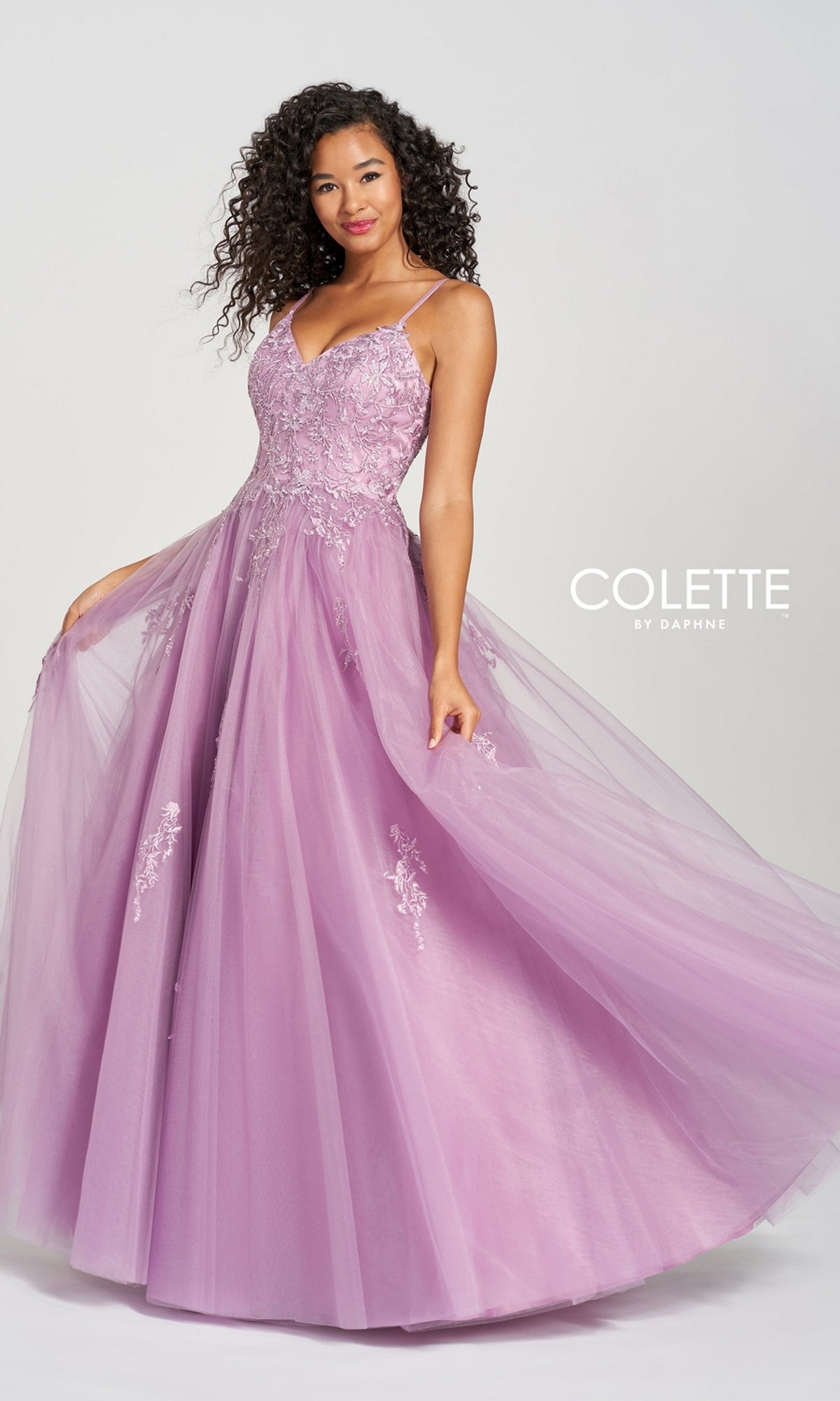 A woman poses against a plain background in the Long Prom Dress CL12212 by Colette—a floor-length lavender tulle ball gown featuring lace, floral embroidery, and thin straps for an elegant touch.