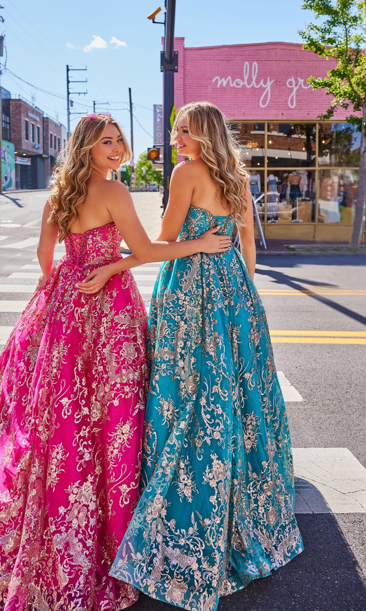 Two women wearing Colette Strapless Long Prom Ball Gown CL5101, one in pink and one in teal, stand arm in arm on a city street in front of the 