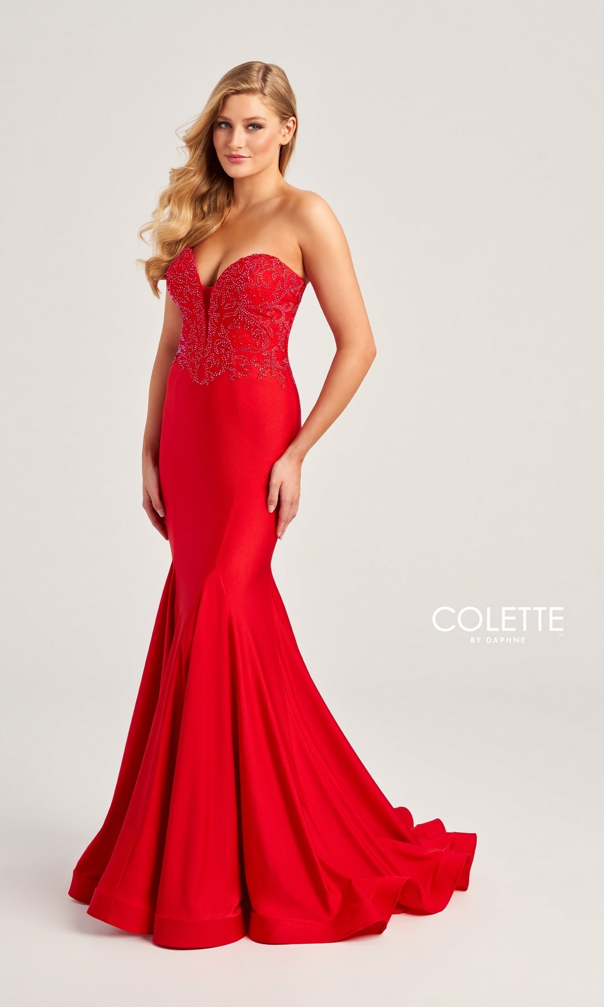 A woman wears the Colette Strapless Long Mermaid Prom Dress CL5112, featuring a fitted red mermaid silhouette and floral beaded bodice, standing against a light background. The 