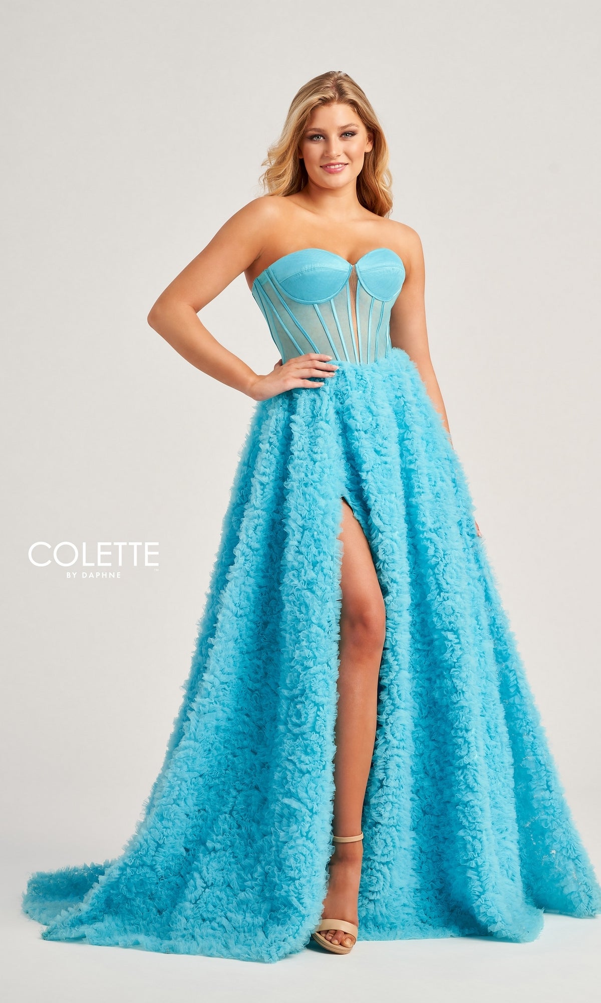 A woman poses in the Bustier Colette Long A-Line Prom Ball Gown CL5114, a sky blue strapless dress with a corset-style bodice and textured, floor-length skirt with a high slit. The background is plain and light-colored.