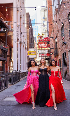 Three women in elegant prom ball gowns, including the Bustier Colette Long A-Line Prom Ball Gown CL5114, walk arm-in-arm down an urban alley lit with string lights and neon signs, smiling at the camera.