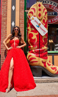 A woman in the Bustier Colette Long A-Line Prom Ball Gown CL5114 and sunglasses poses beside a large decorative cowboy boot outside a storefront.