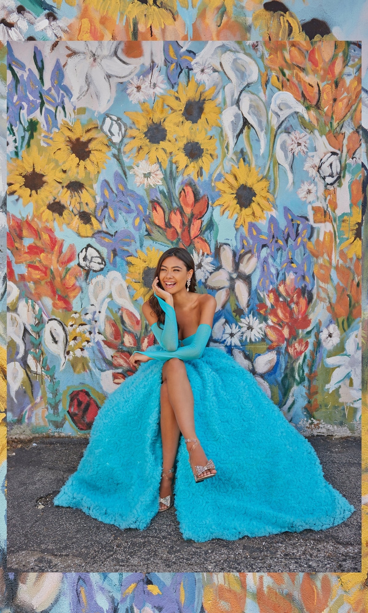 A woman in a bright blue Bustier Colette Long A-Line Prom Ball Gown CL5114 sits smiling before a colorful floral mural, her stunning dress harmonizing beautifully with the vivid background.