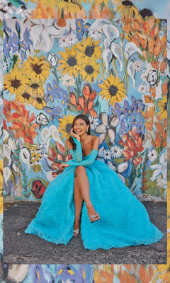 A woman in a bright blue Bustier Colette Long A-Line Prom Ball Gown CL5114 sits smiling before a colorful floral mural, her stunning dress harmonizing beautifully with the vivid background.