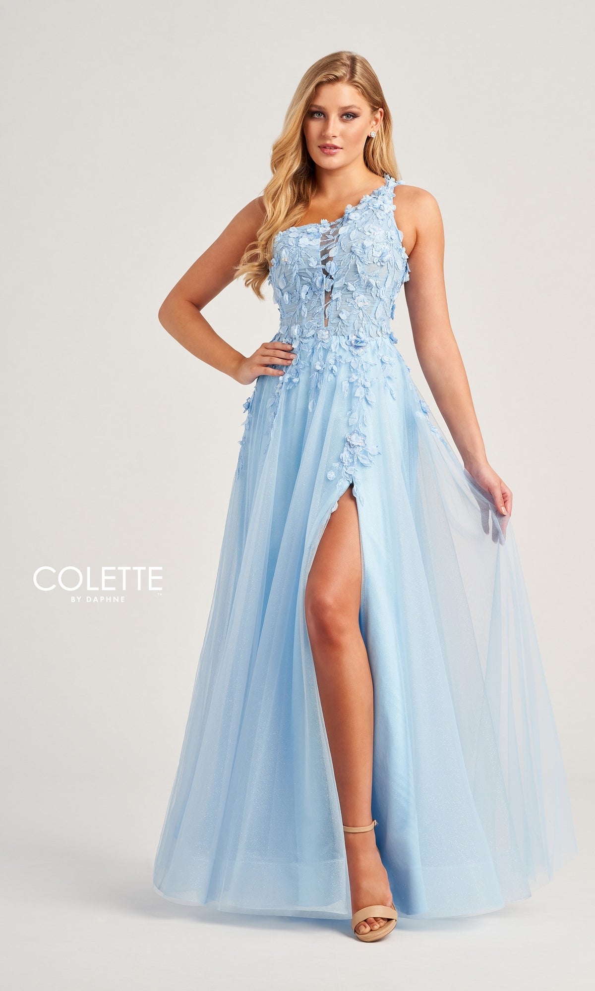 A woman wears the Colette One-Shoulder Designer Prom Dress CL5124 in light blue, featuring floral embroidery, a sheer tulle skirt, and a high leg slit, standing against a plain background.