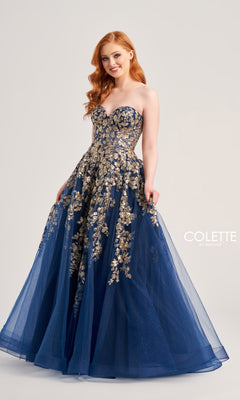 A woman wearing the Colette Strapless Sweetheart Prom Ball Gown CL5136, which has gold floral embroidery and sparkly lace appliqué on a full navy blue tulle skirt, stands against a plain background.