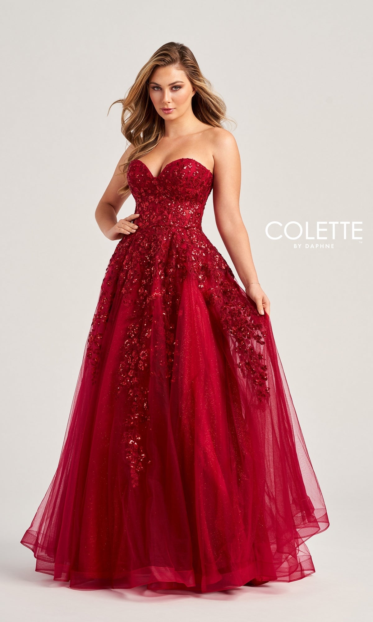 A woman wears the Colette Strapless Sweetheart Prom Ball Gown CL5136—a floor-length red gown with sparkly lace appliqué, a fitted bodice, and a full tulle skirt that creates an elegant silhouette against a plain background.