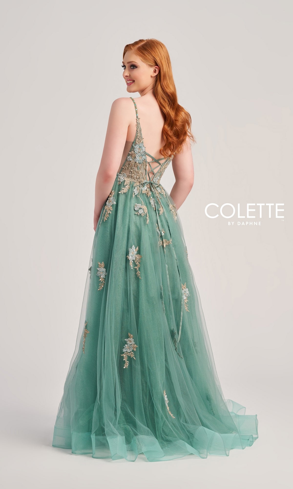 A woman stands facing away in a studio wearing the Glitter-Lace Colette Designer Prom Gown CL5143—a green tulle evening dress featuring floral lace embroidery and a lace-up back.