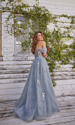 A woman in an off-shoulder, light blue Colette Off-the-Shoulder Lace Prom Dress CL5169 smiles in front of a weathered white wooden wall with climbing green vines.