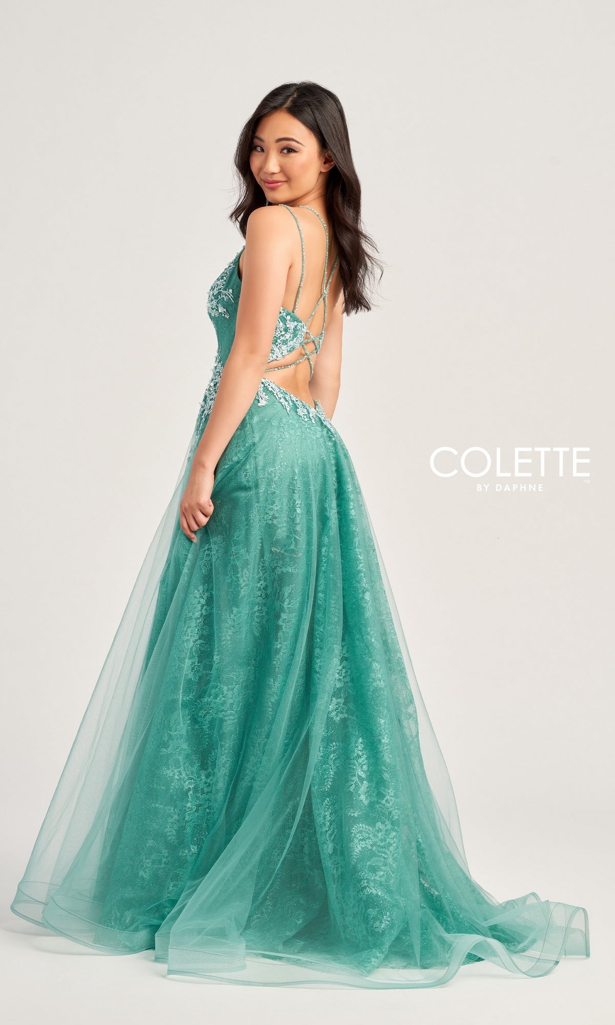 A woman in the Glitter-Lace Colette Long Prom Ball Gown CL5197—teal, floor-length, A-line with beaded-lace floral appliqué and open back—stands looking over her shoulder against a light background. 