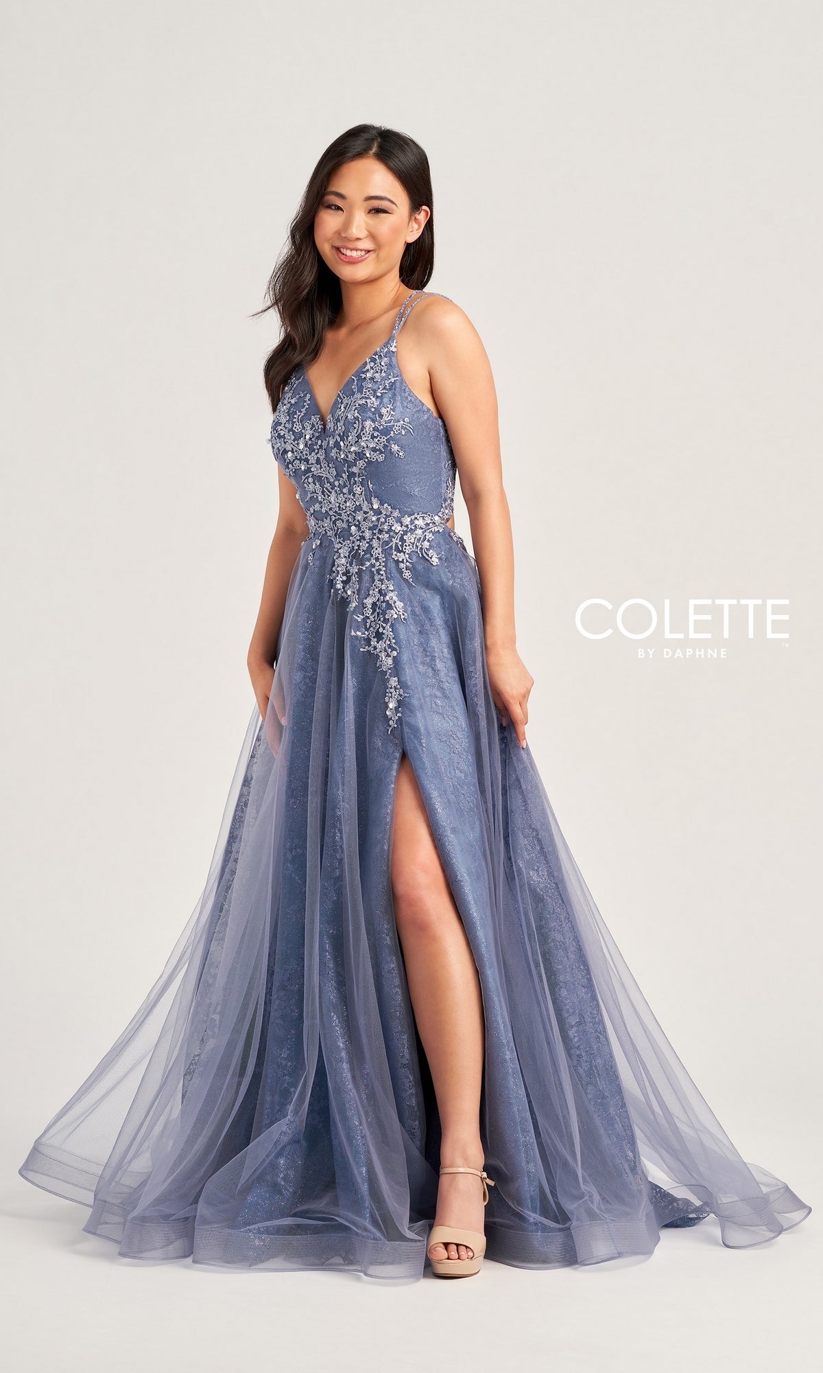 A woman smiles in a sleeveless, floor-length blue Glitter-Lace Colette Long Prom Ball Gown CL5197 with beaded floral appliqué and a thigh-high slit. The text 
