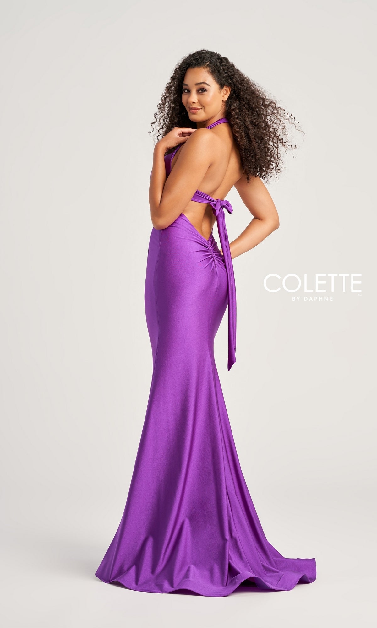 A woman with curly hair models the Colette Front-Knot Long Halter Prom Dress CL5199—an elegant, floor-length, purple spandex jersey gown featuring a halter neck, side bow, and backless design. 