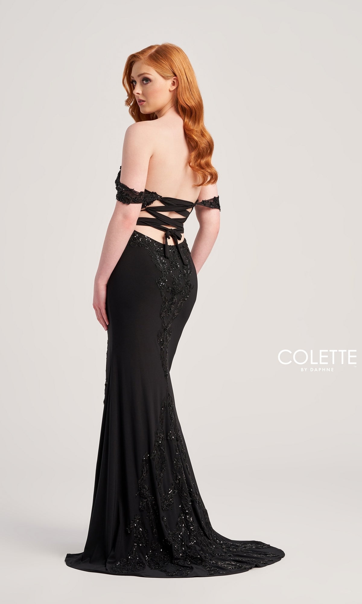 A woman with long red hair wears the Colette Off-Shoulder Long Formal Prom Dress CL5276, a black, floor-length gown with beaded lace applique, sequin embellishments, an off-shoulder neckline, and a lace-up back against a plain background.