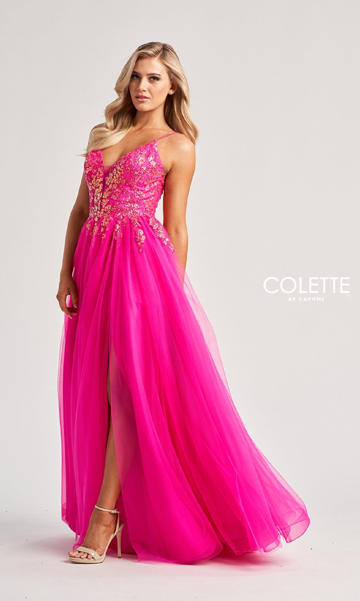 A woman poses in the Colette CL8030—a vibrant pink long prom dress with a sequined bodice, gold embellishments, a floor-length slit tulle A-line skirt, and elegant details—against a plain background.