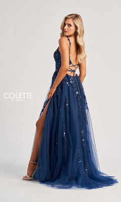 A woman stands in profile against a plain light background wearing the Colette CL8060, a dark blue, floor-length prom dress with strappy design, sequin details, and a side slit.
