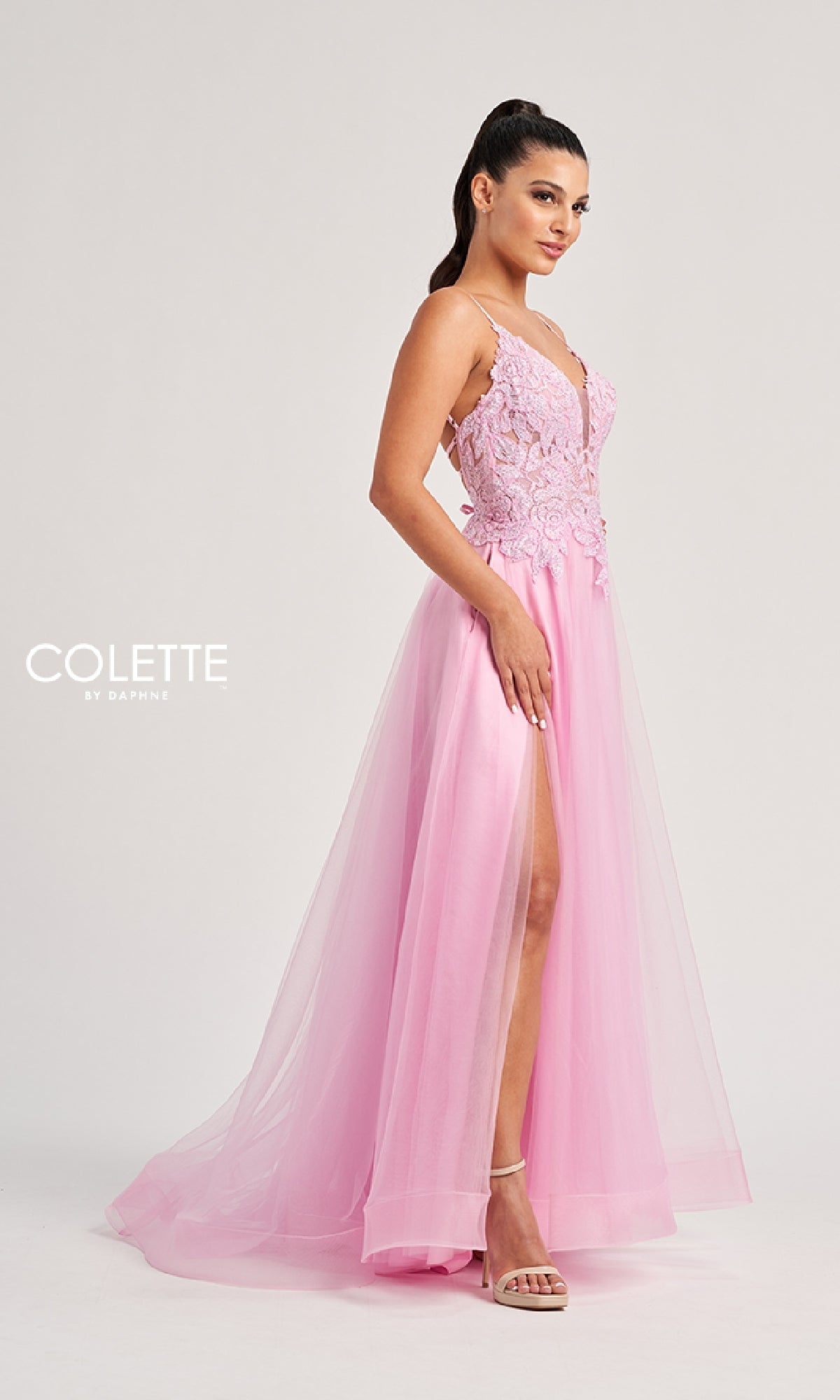 Woman in a pink lace and tulle A-line Long Prom Dress: Colette CL8100 with a high slit, standing in heels against a plain background. Delicate lace embroidery enhances the elegant look. The 
