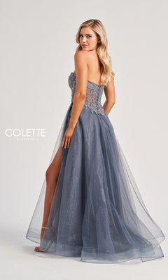 A woman in the Colette CL8110 long prom dress stands sideways, showcasing its strapless embroidered corset bodice, sheer overlay, floor-length blue a-line skirt, and high slit against a plain background.