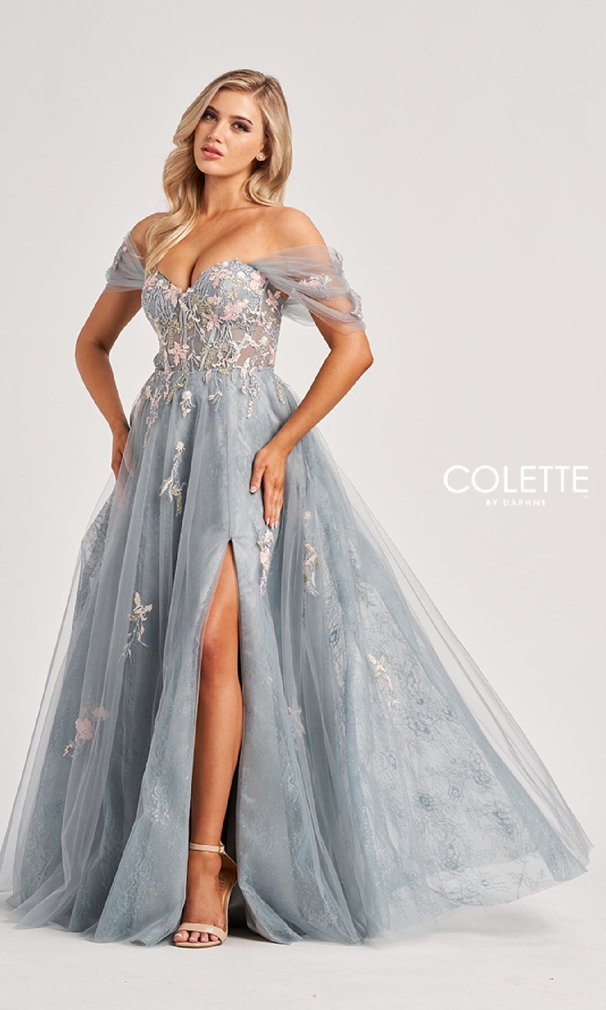 A woman models the Colette CL8150 long prom dress—an off-the-shoulder blue tulle gown with a sweetheart neckline, floral embroidery, sheer sleeves, and thigh-high slit—standing before a plain light background.