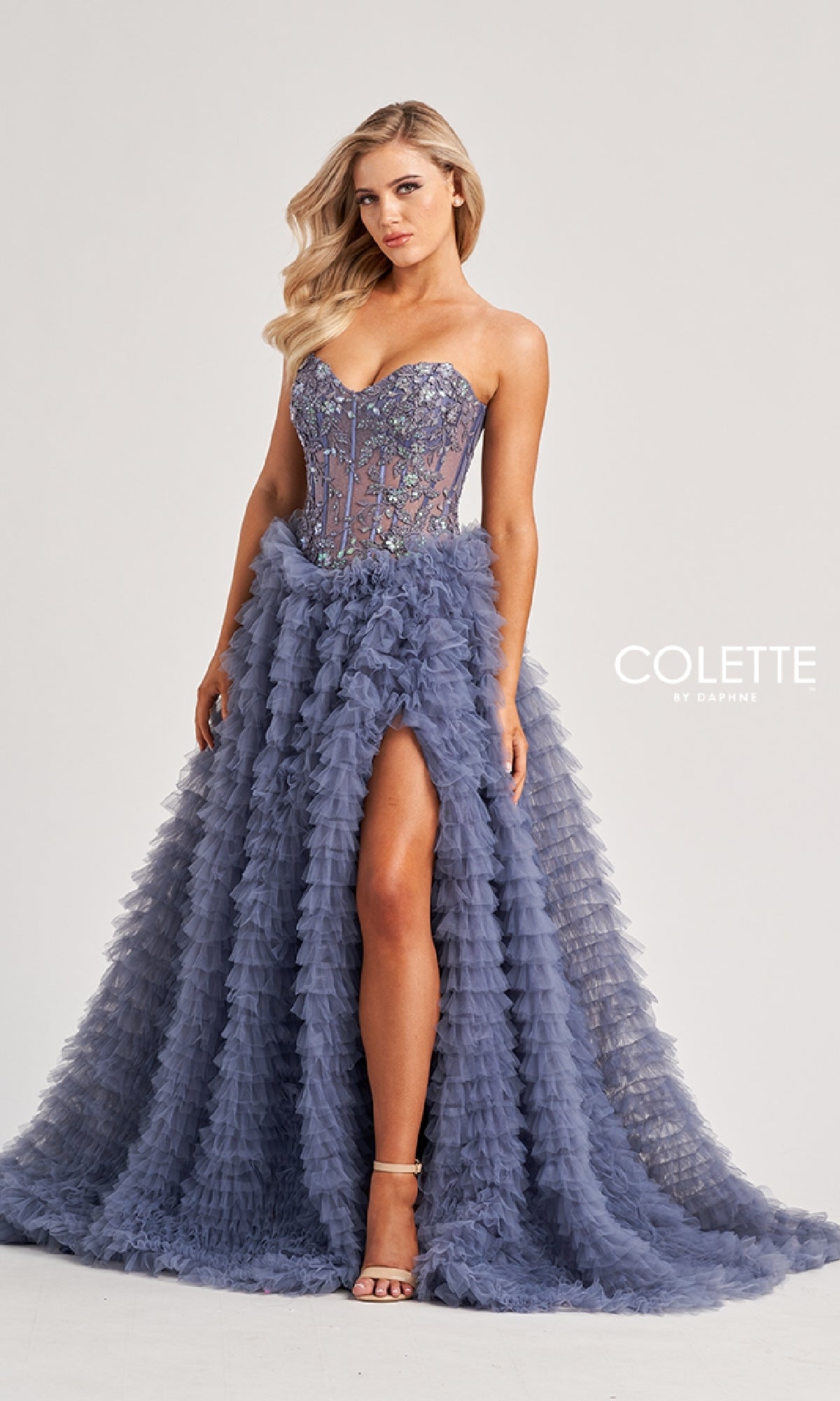 A woman stands against a plain background wearing the Colette CL8170 long prom dress—a strapless, smokey blue lace corset gown with a ruffled tulle skirt and high slit, radiating ball-gown elegance.