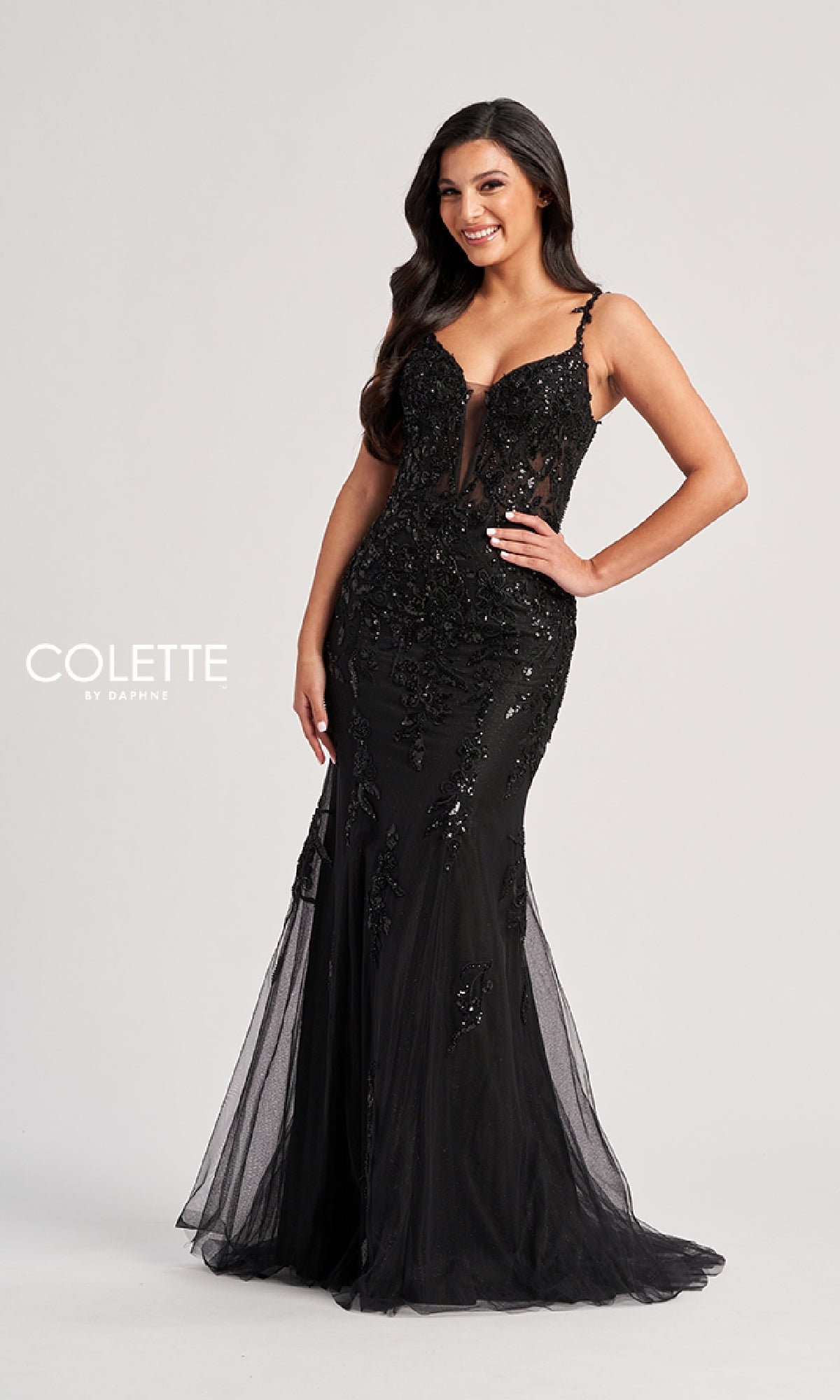 A woman smiles with one hand on her hip, wearing the Colette CL8210 Long Prom Dress—a fitted black lace evening gown with a sheer bodice and mermaid silhouette—against a plain light background.