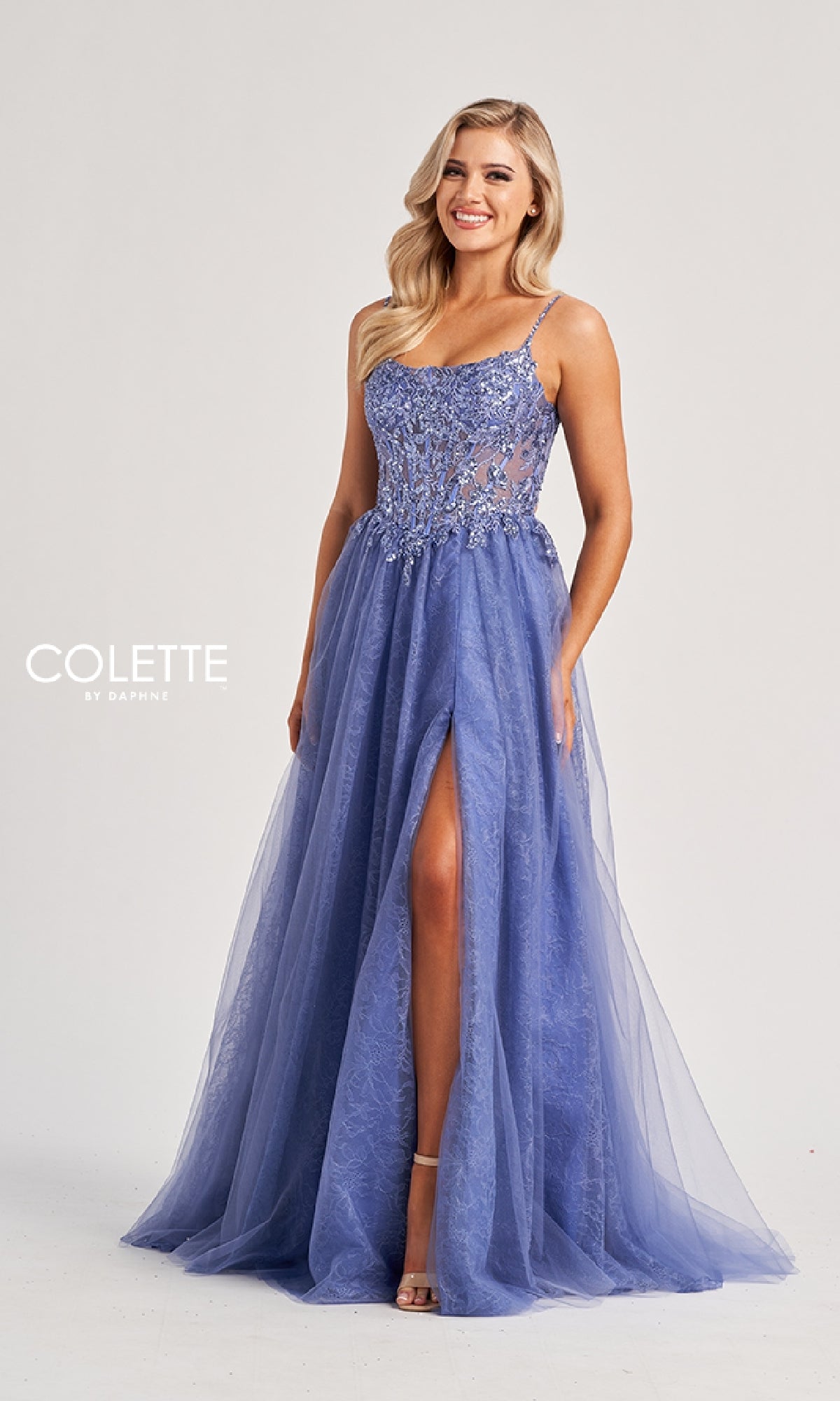 A woman wears the Colette CL8240 long prom dress, a sleeveless lavender gown with floral embellishments, an embroidered bodice, high slit, and a flowing tulle skirt.