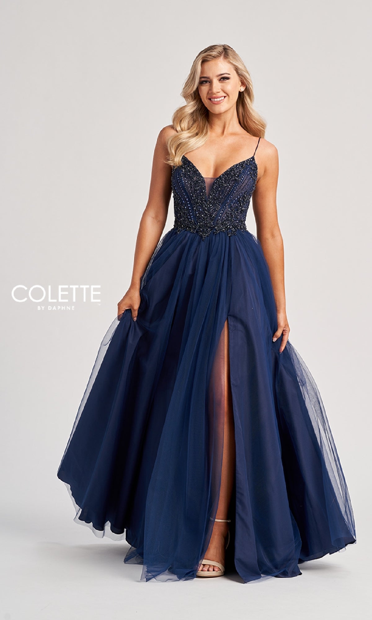 A woman models the Long Prom Dress: Colette CL8250, a navy blue gown with a sequin bodice, thin straps, and high slit, standing against a plain light background.