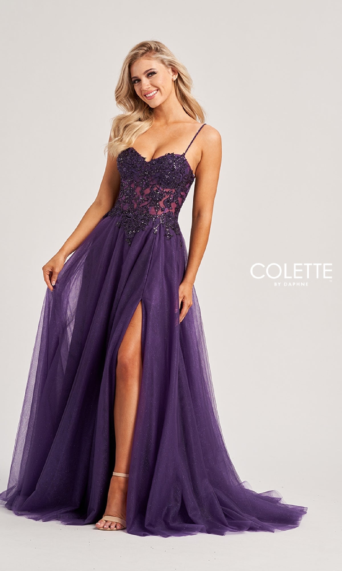 A woman stands and smiles against a plain background, wearing the Colette CL8280 long prom dress—a floor-length purple A-line gown with thin straps, lace bodice, and high slit.