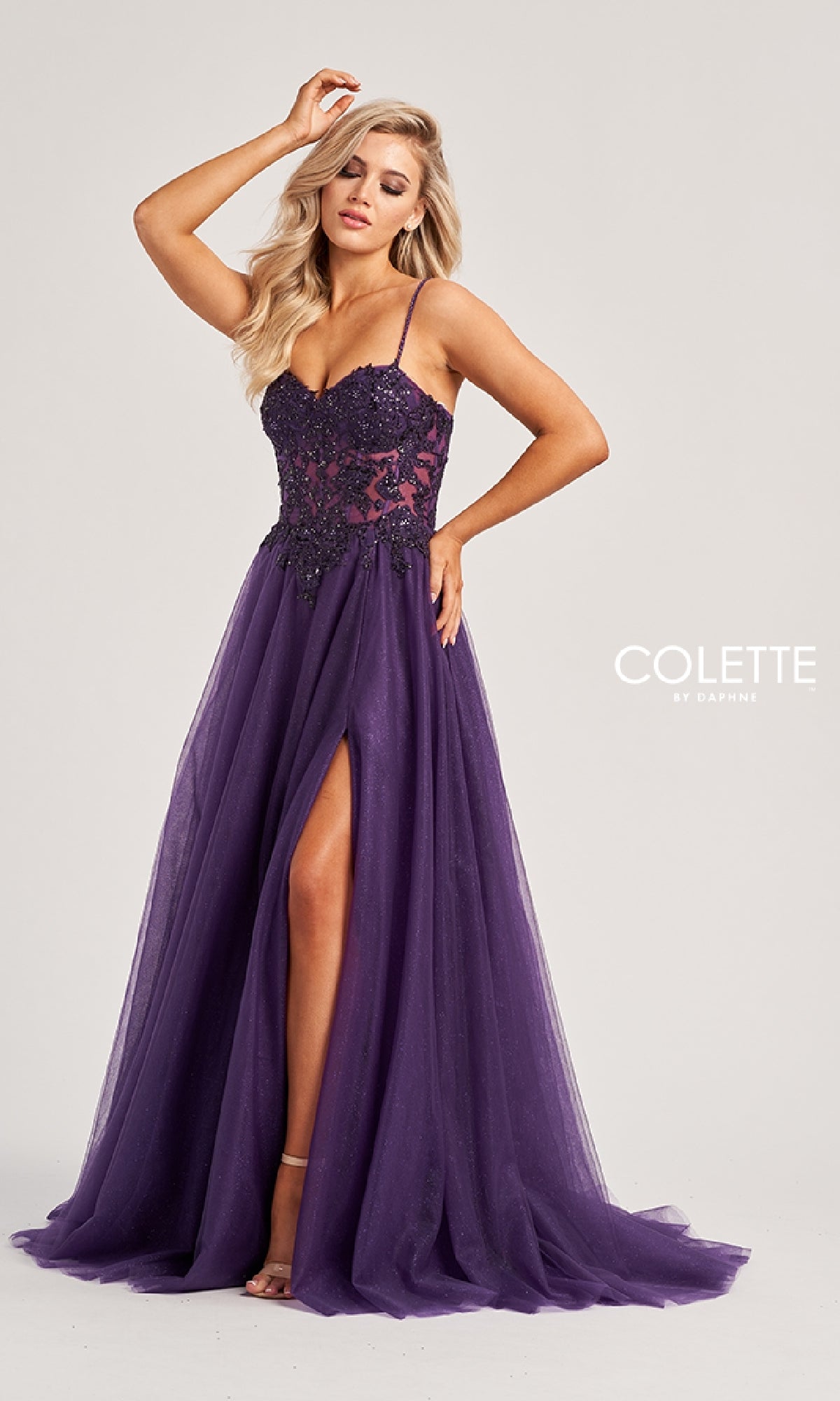 A woman models the Colette CL8280, a long purple prom dress with a floor-length sheer overlay, sleeveless cut, floral bodice detailing, and high front slit, posed against a plain background.
