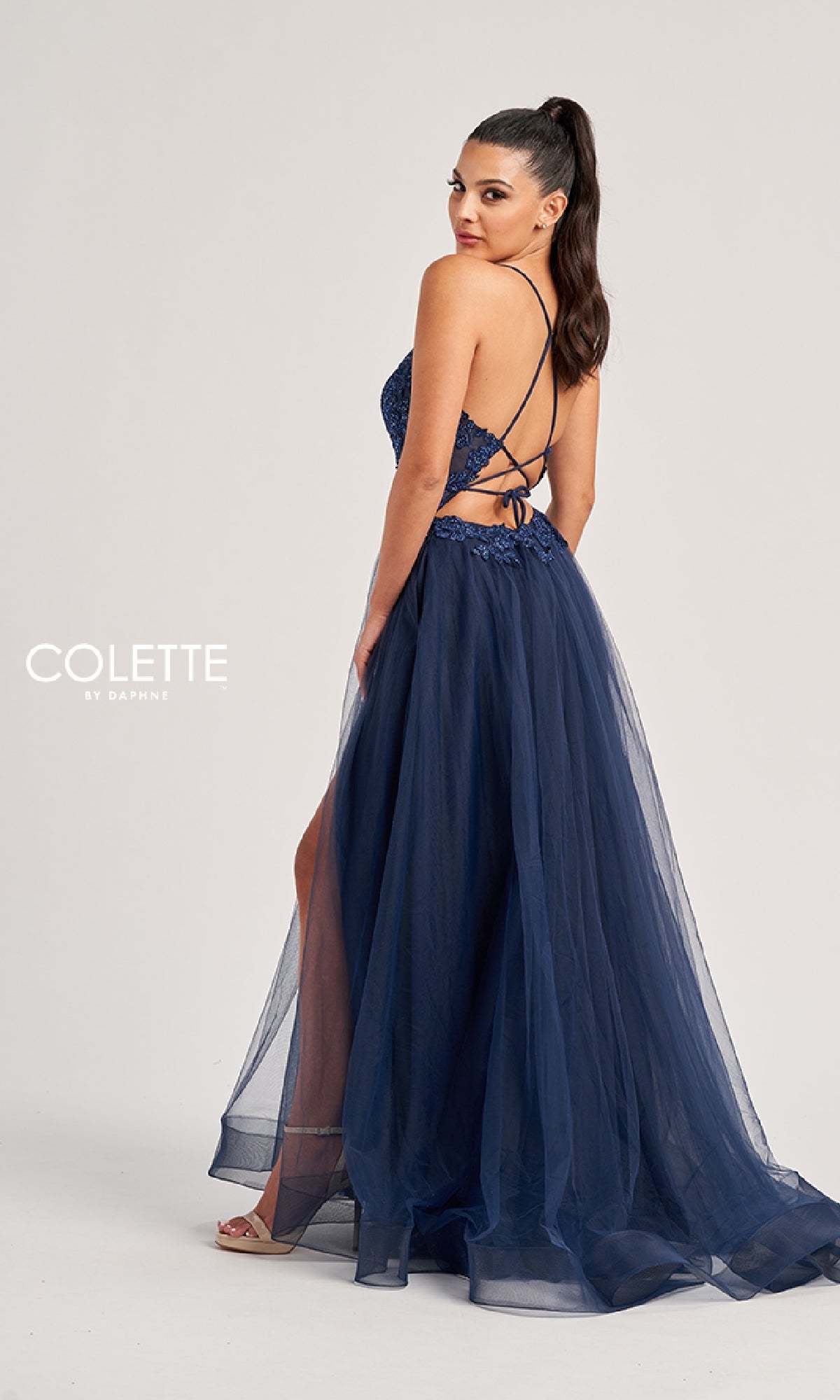 Woman in the Colette CL8320 long prom dress—navy blue, floor-length with a high slit, crisscross back straps, and an embroidered bodice—stands looking over her shoulder. 