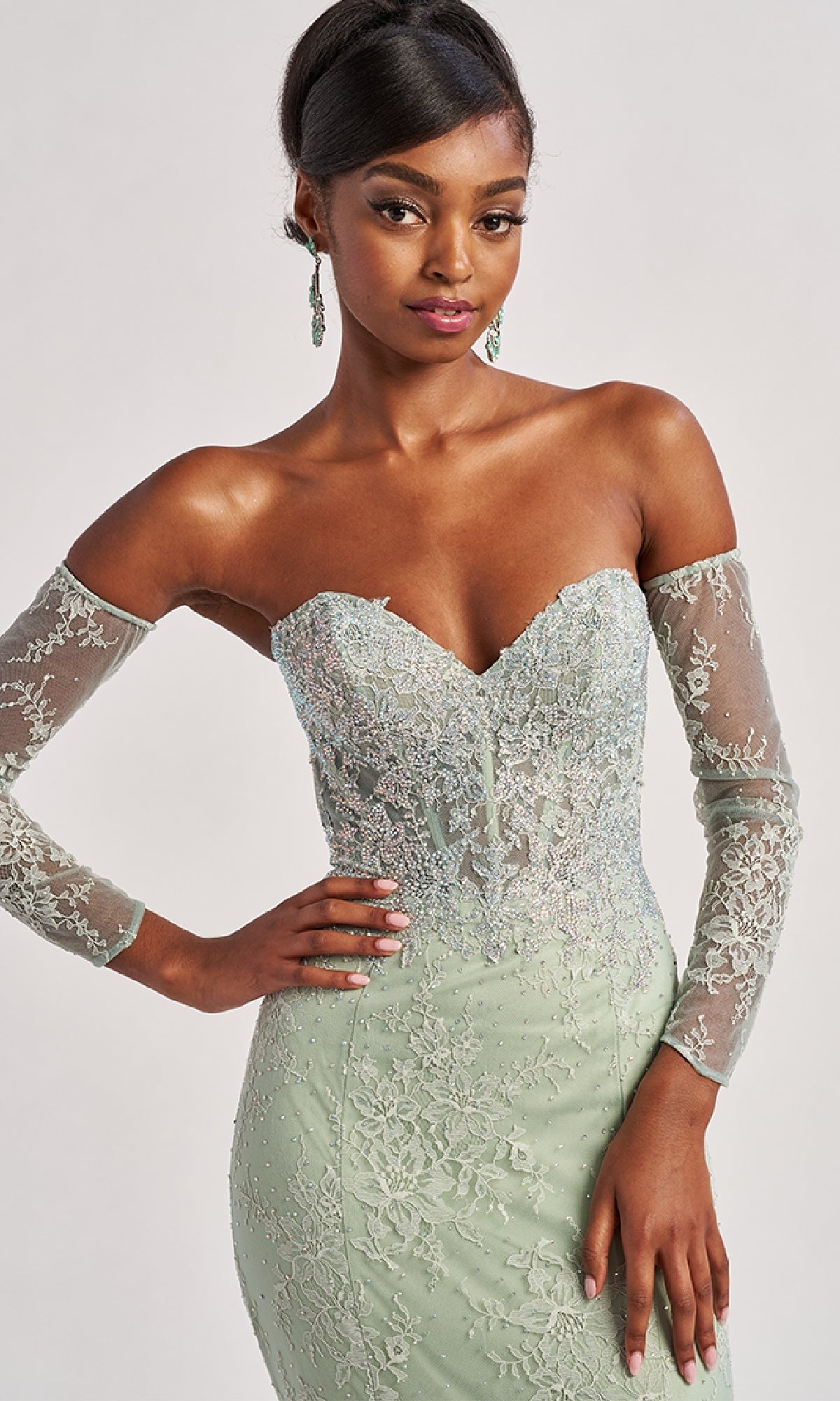 Woman wearing the Colette CL8415 Strapless Long Lace Prom Dress in light green, featuring sheer sleeves and embroidered details, poses with one hand on her hip.