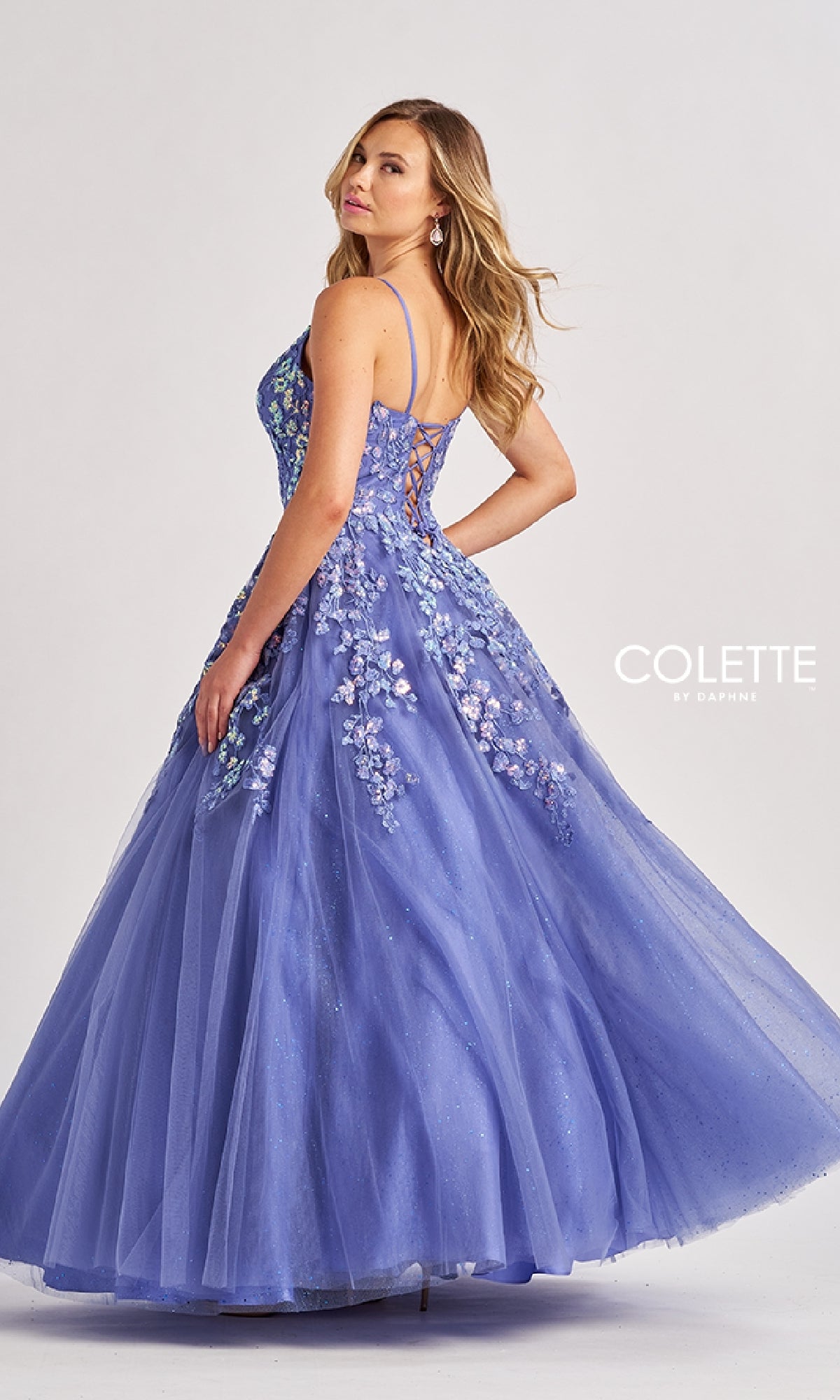 A woman wears the Low V-Neck Glitter Prom Ball Gown: Colette CL8420, a lavender dress featuring floral embellishments and a lace-up back, as she stands against a plain background.