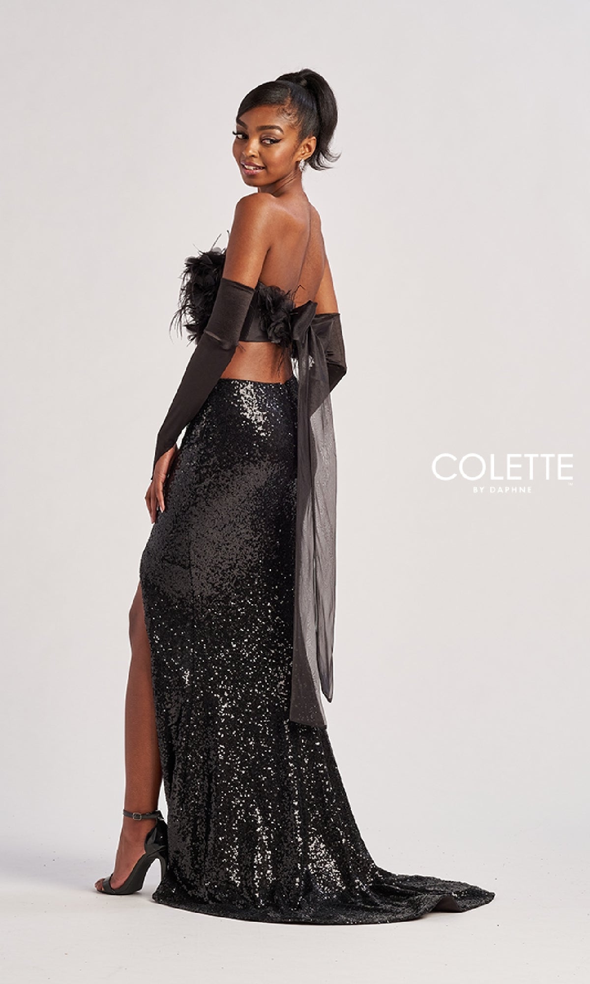 A woman poses in the Colette CL8450 long prom dress, a strapless two-piece featuring a feathered crop top and sequined floor-length skirt. She accessorizes with black gloves and heels, creating a chic look against a simple light background.