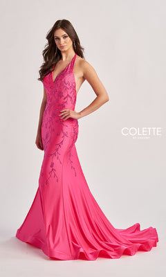 A woman stands against a plain background wearing the Colette CL8455, a floor-length bright pink halter prom dress with embroidery, a v-neckline, fitted bodice, flared skirt, and a small train.