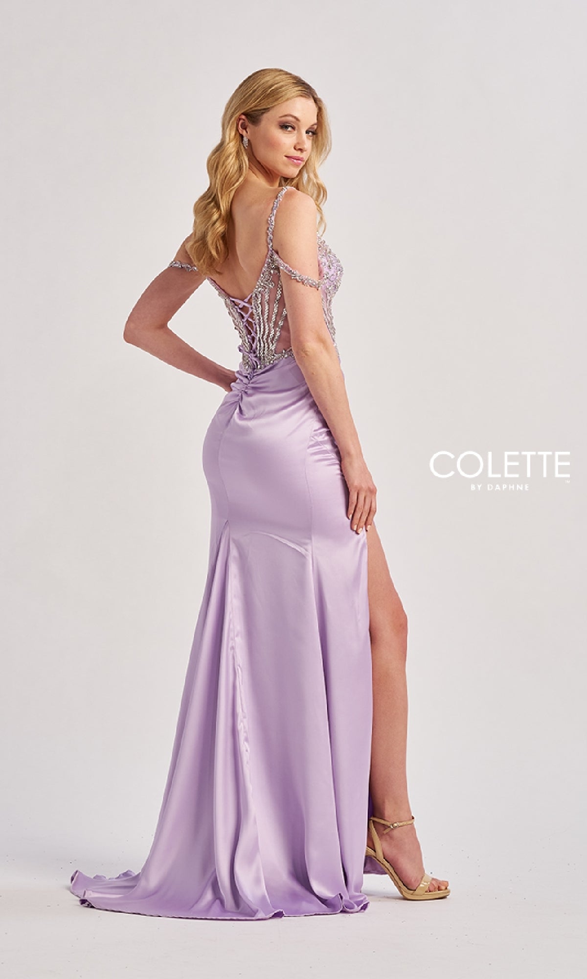 A woman in the Colette CL8460 long prom dress, featuring a lilac hue, sheer embroidered corset bodice, and thigh-high slit, poses sideways against a plain background. The text 