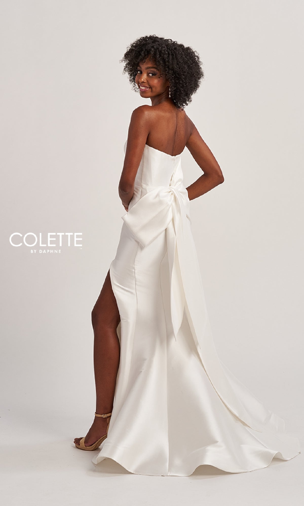 A woman in the Colette CL8470 Back Bow Strapless Long Prom Dress, featuring a large back bow and side slit, stands facing away, looking over her shoulder and smiling.