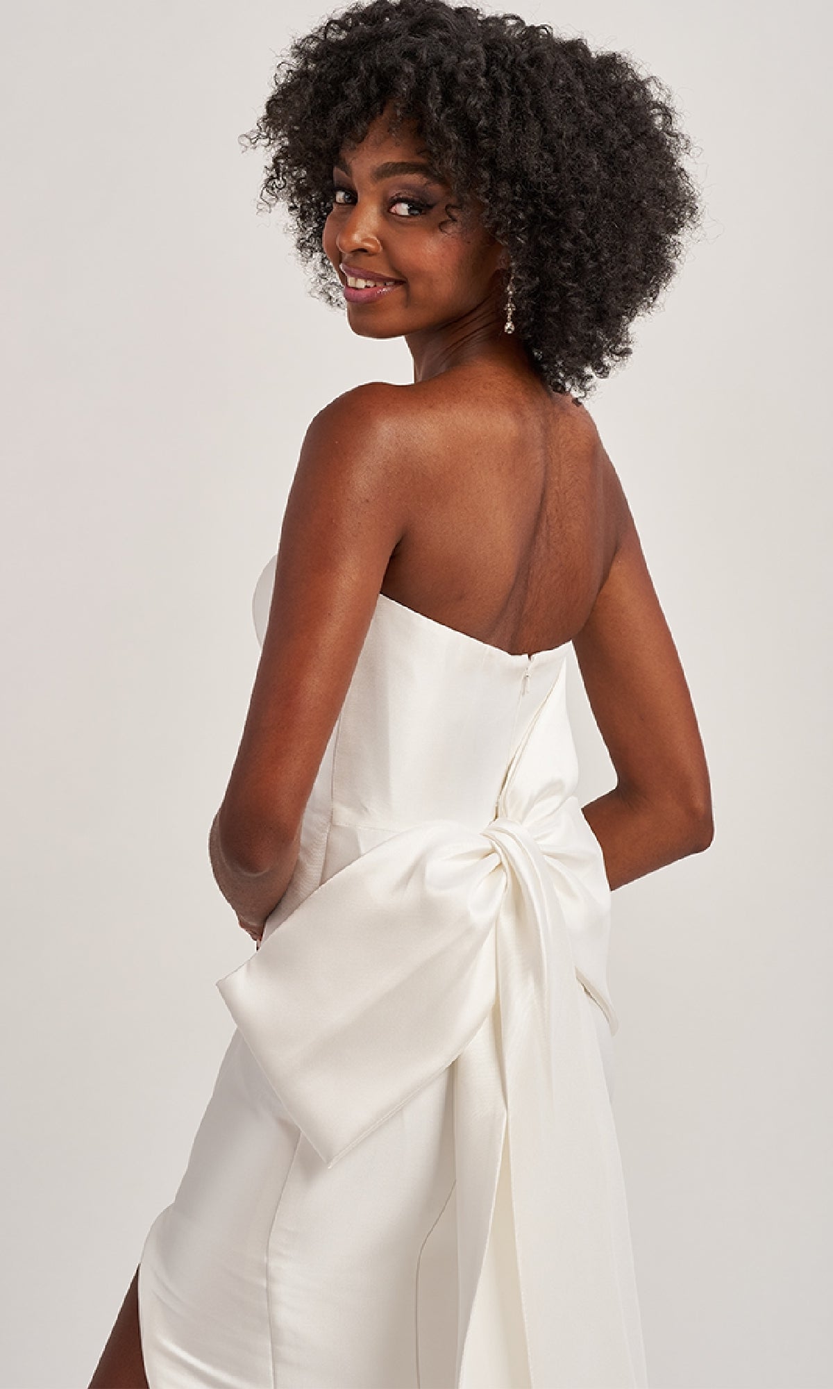 Model wears the Colette CL8470 Back Bow Strapless Long Prom Dress, posing and smiling over her shoulder to showcase the statement bow against a plain background.