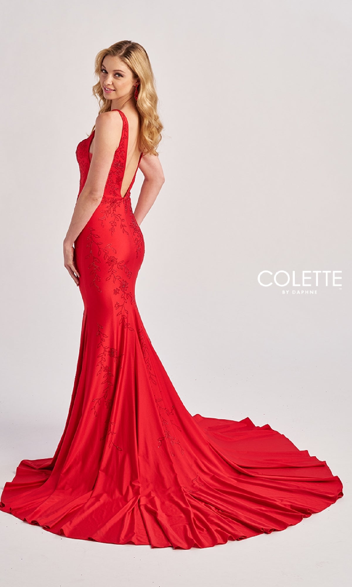 A woman poses in front of a plain background wearing the Colette CL8475, a floor-length fitted red mermaid prom dress with lace detailing and a train. The 