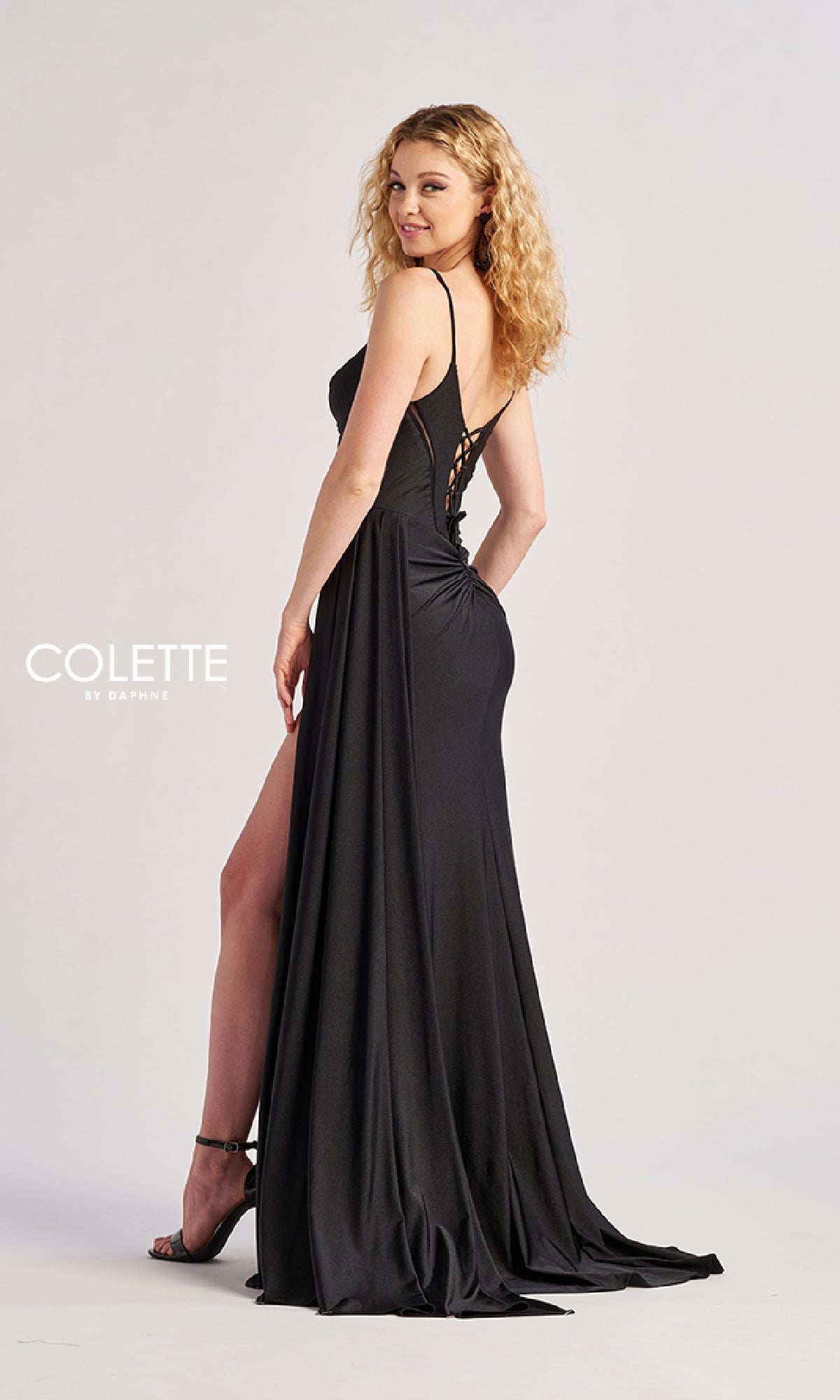 Woman in the Sheer-Cut-Out Long Prom Dress: Colette CL8480, a black gown featuring a thigh-high slit and open back, standing in profile and looking over her shoulder against a plain background.